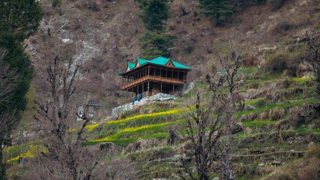 A peaceful rural scene in Kasol with a traditional wooden house amidst trees and terraced hills.